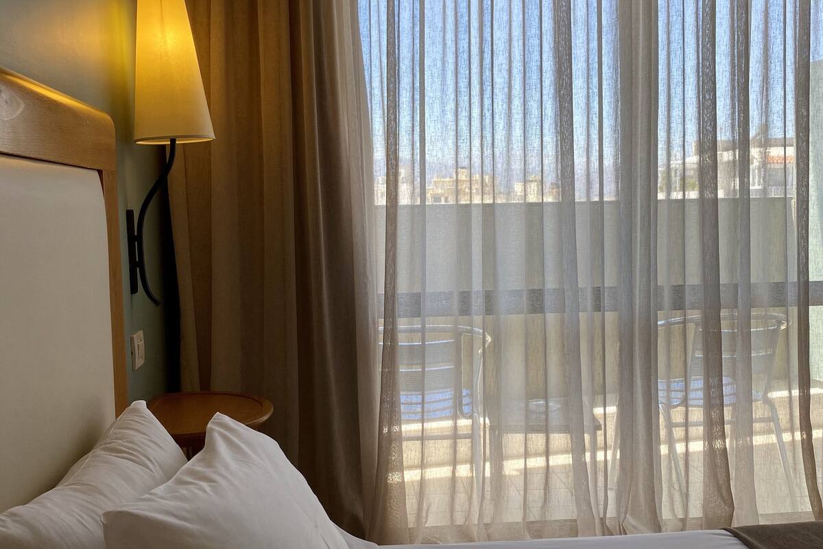 A hotel room with a bed, white pillows, a lamp, and a view of the outdoors through sheer curtains.
