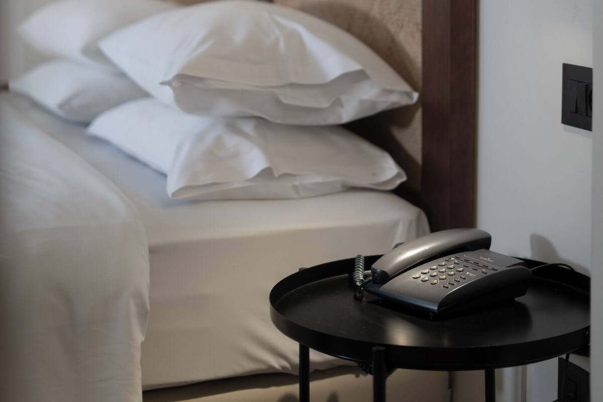 A hotel room with a bed, pillows, a bedside table with a telephone, and wall-mounted light fixtures.