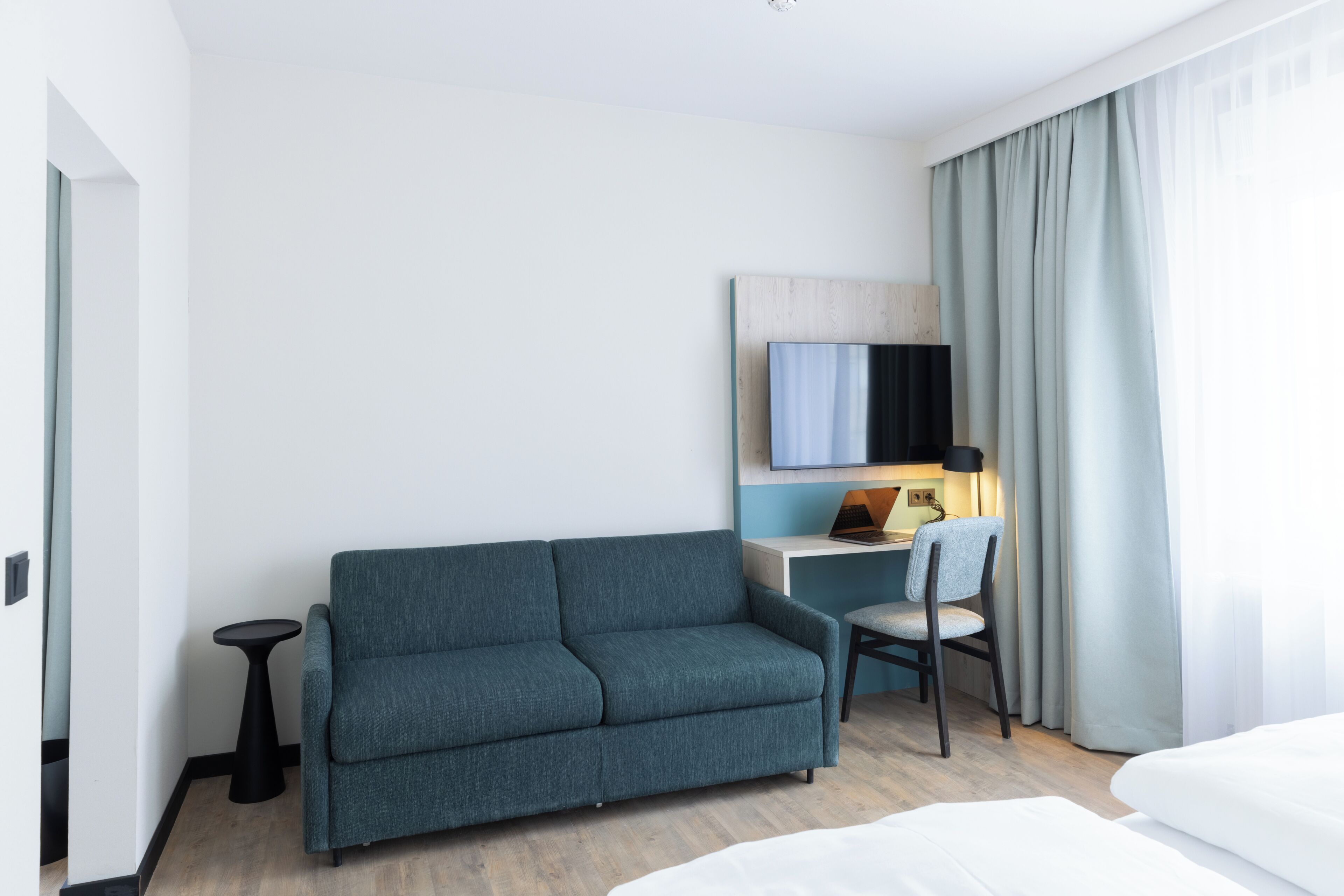 A modern hotel room with a sofa, a desk with a chair, a television, and a bed.