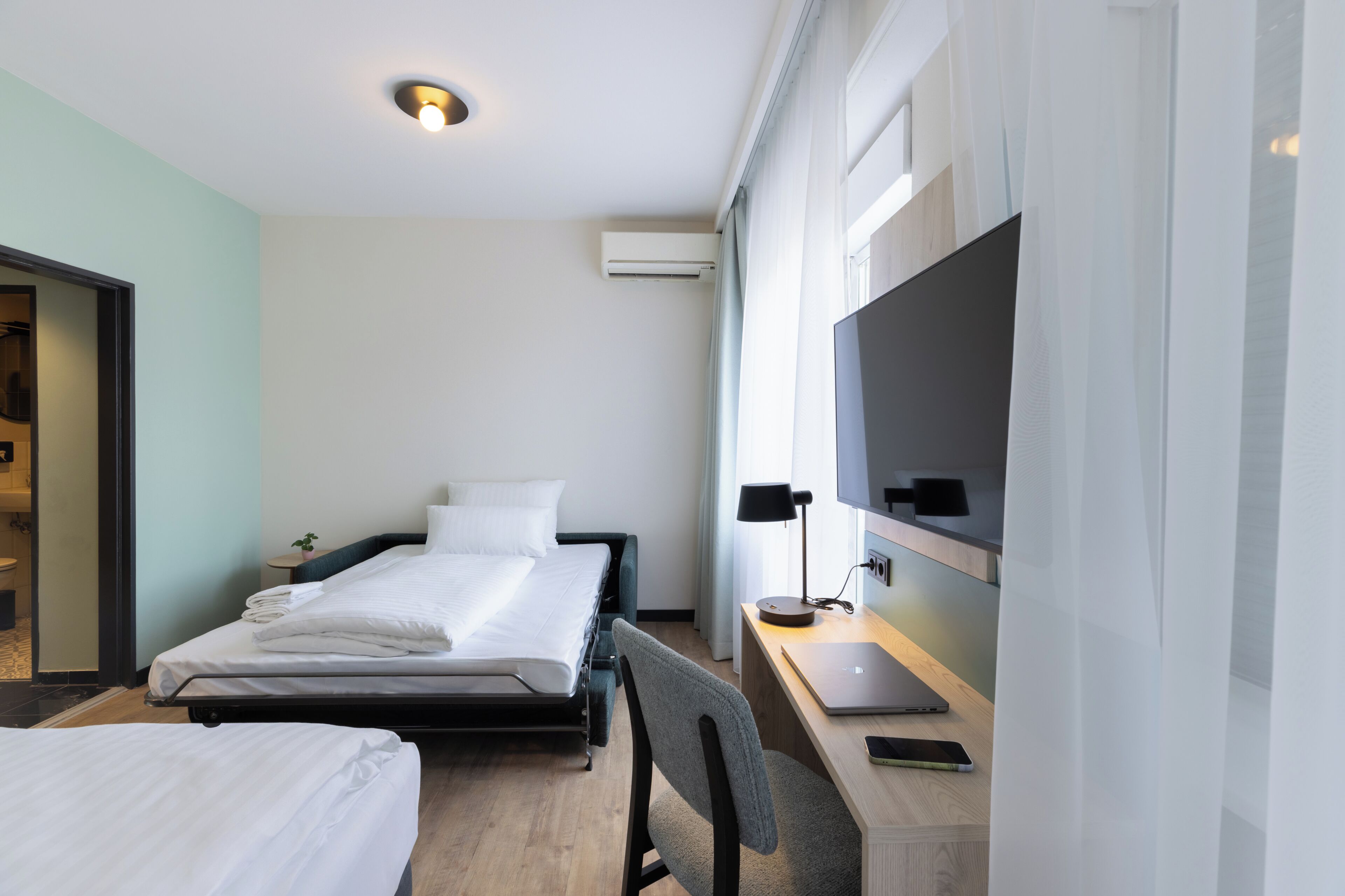 A hotel room with a bed, a desk with a laptop, a chair, a TV, and an air conditioning unit.