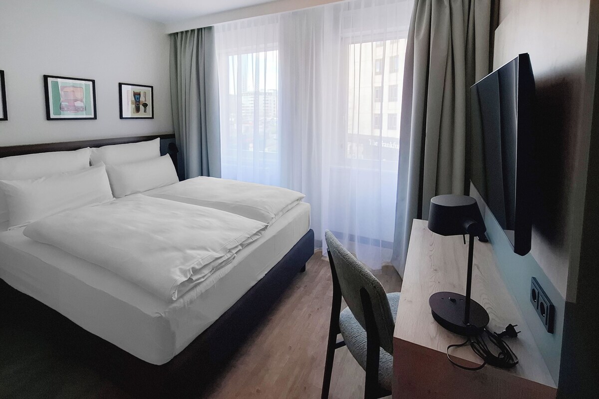 A hotel room with a bed, a desk with a lamp, a chair, a TV, and a window with curtains.