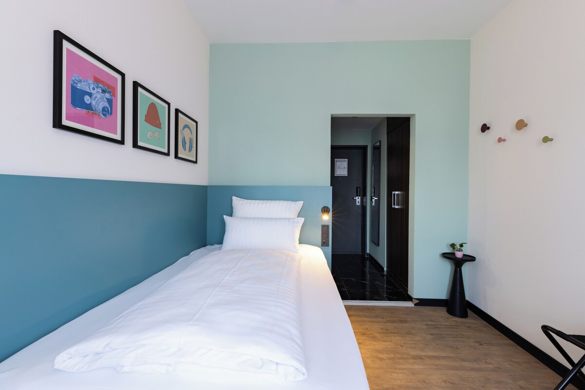 A hotel room with a bed, a wall-mounted camera, a small table with a plant, and a door leading to a bathroom.