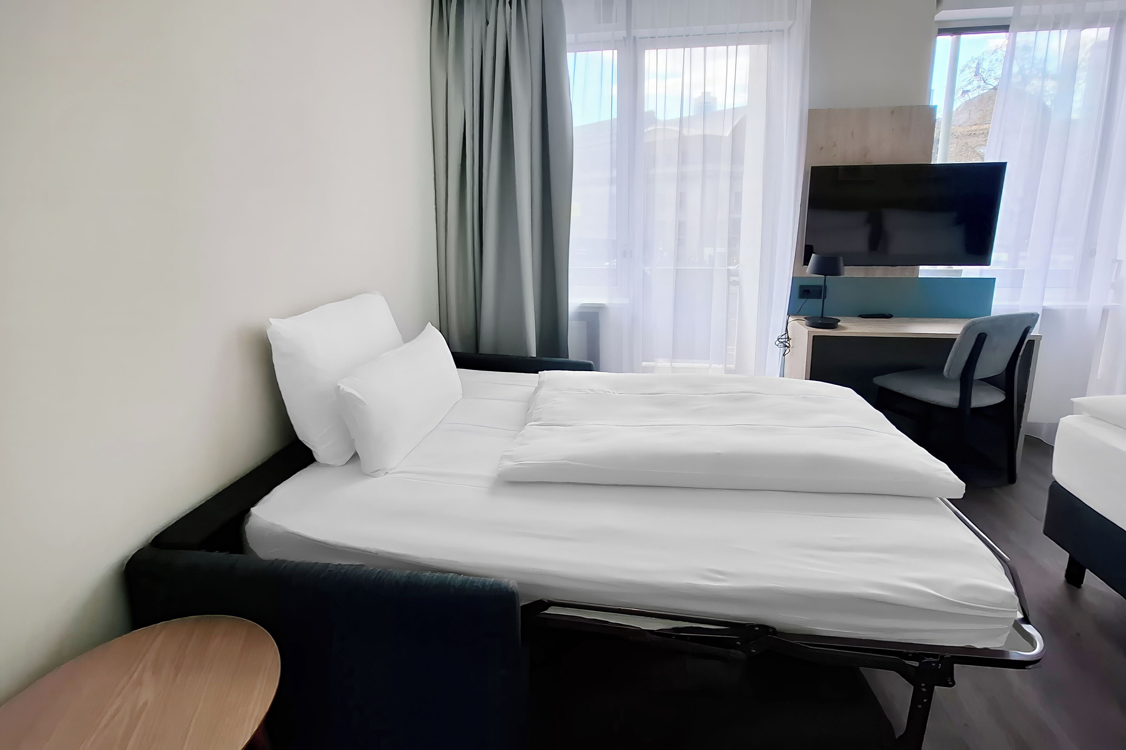 A hotel room with a bed, a desk with a computer, a TV, and a window with curtains.