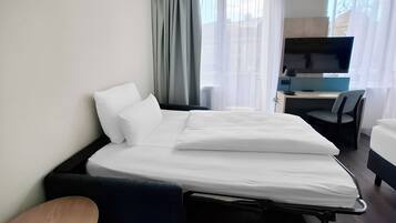 A hotel room with a bed, a desk with a computer, a TV, and a window with curtains.