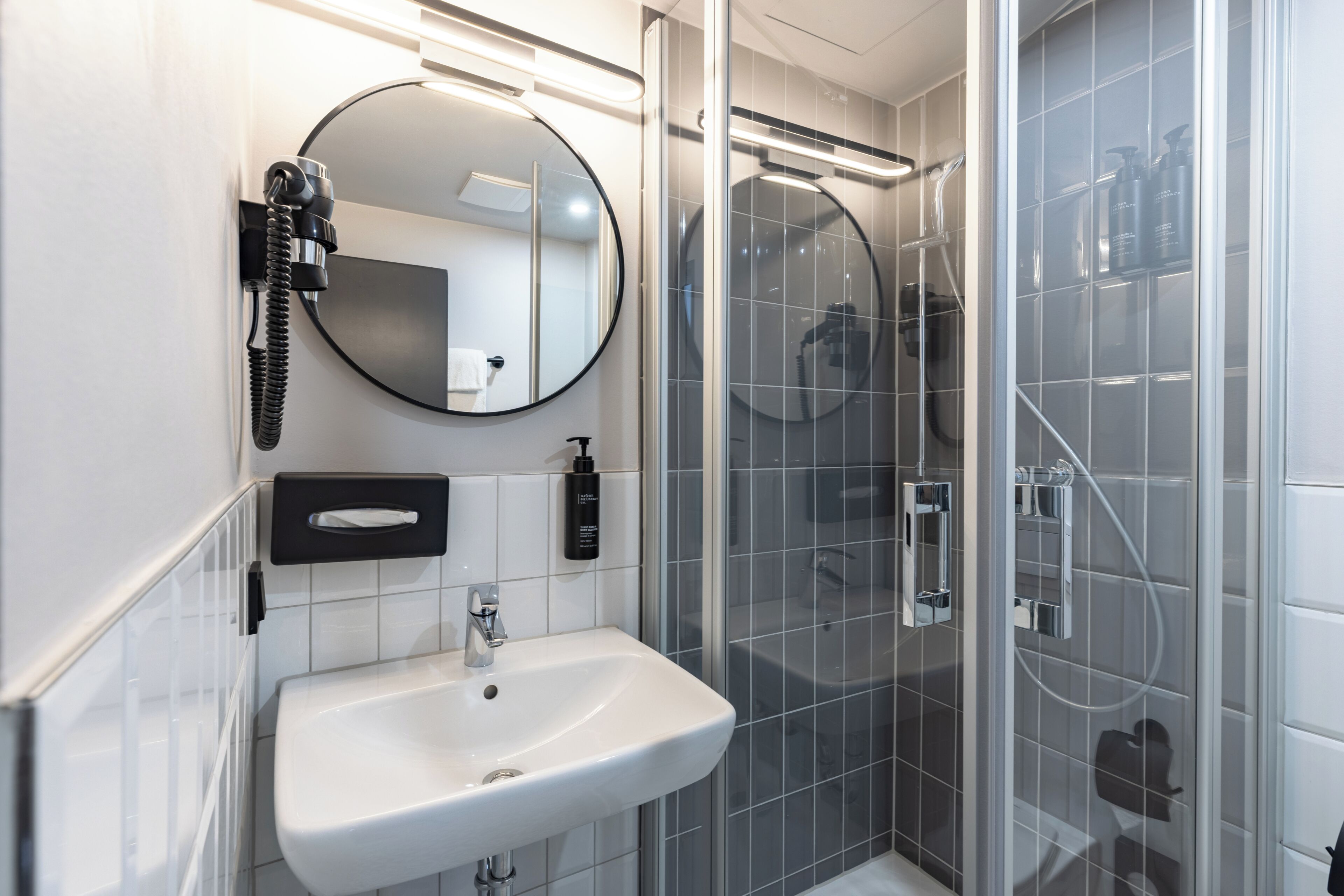Ein modernes Badezimmer mit einem weißen Waschbecken, einem runden Spiegel, einer Handbrause und einer Duschkabine aus Glas.