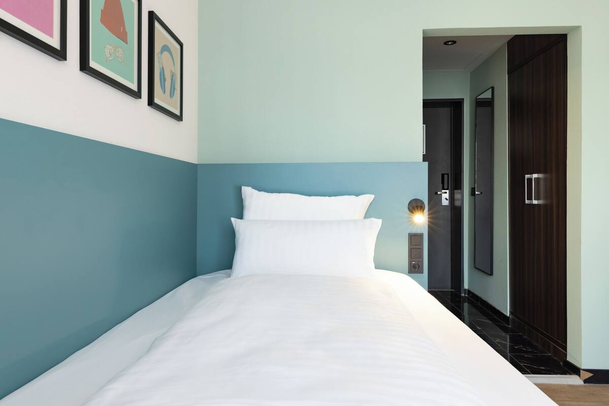 A neatly made bed in a room with teal walls, a door, and a framed picture on the wall.