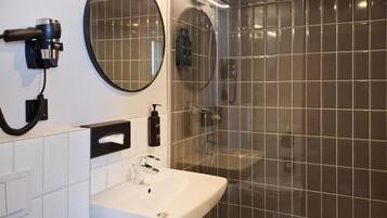 A bathroom with a white sink, a round mirror, a hairdryer, and a shower area with tiled walls.