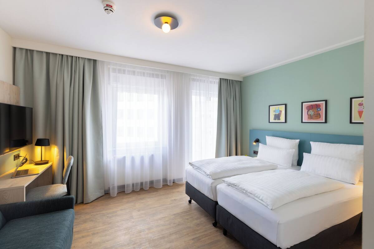 A hotel room with two beds, a desk with a lamp, a TV, and a window with curtains.