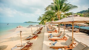 Une plage bordée de rangées de transats sous des parasols, une piscine et des palmiers.