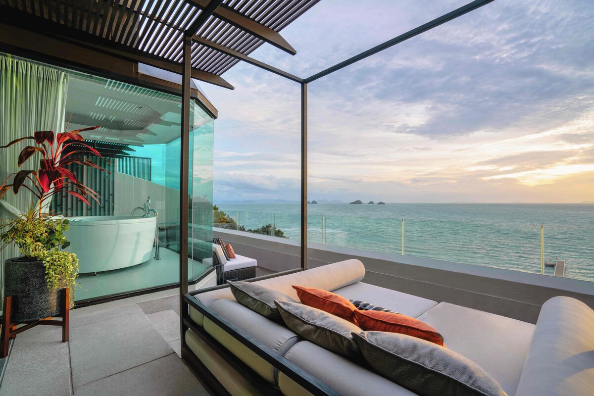 Un balcon moderne avec une paroi en verre, un canapé avec des coussins, une plante en pot et une vue sur la mer.