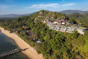 An aerial view of a coastal resort with multiple buildings, a beach, and a forested hillside.
