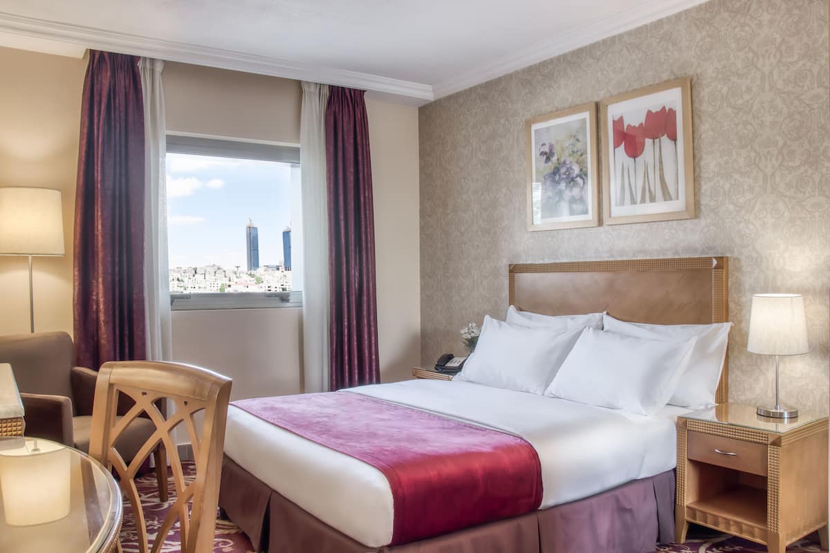 A hotel room with a bed, a chair, a desk, and a window with a city view.