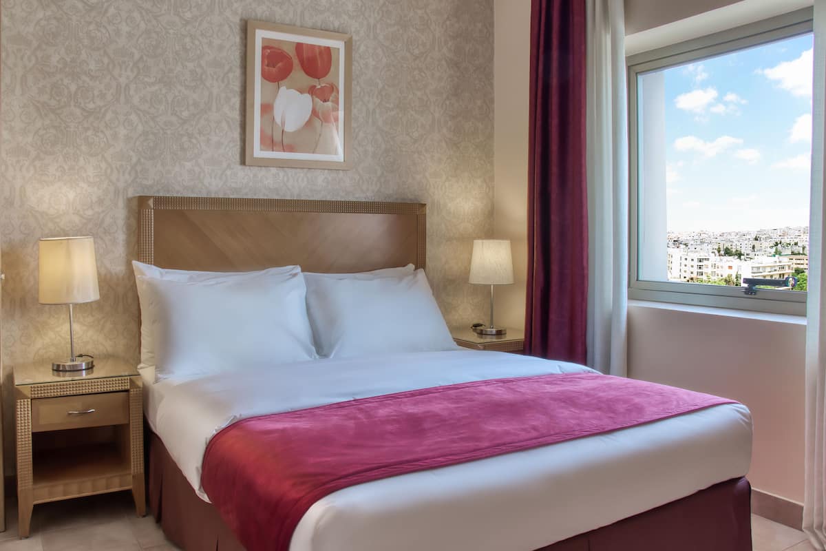 A hotel room with a large bed, two bedside tables with lamps, a view of the city through a window, and a framed picture on the wall.
