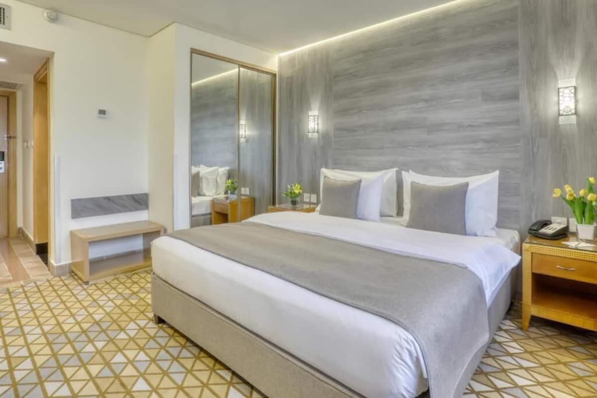 A hotel room with a large bed, a bedside table with a lamp, a mirror, and a small bench.