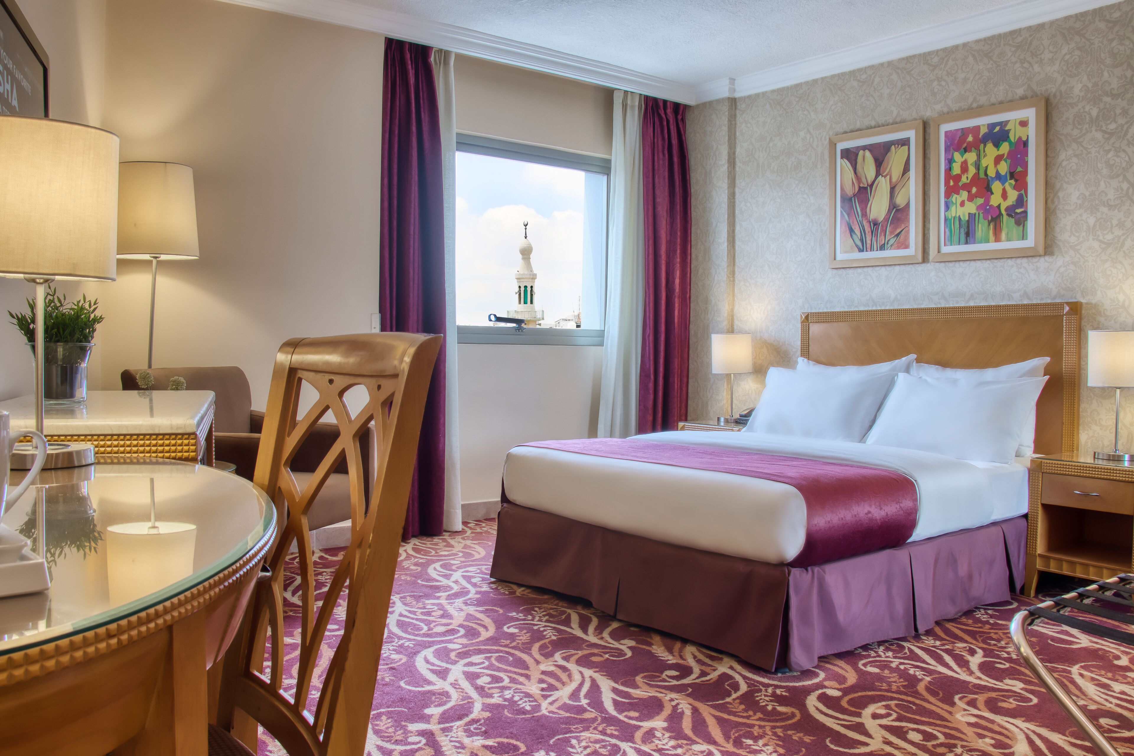 A hotel room with a bed, a desk with a lamp, a chair, a window with curtains, and two framed pictures on the wall.