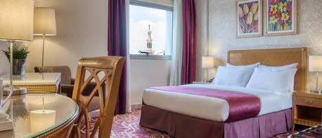 A hotel room with a bed, a desk with a lamp, a chair, a window with curtains, and two framed pictures on the wall.
