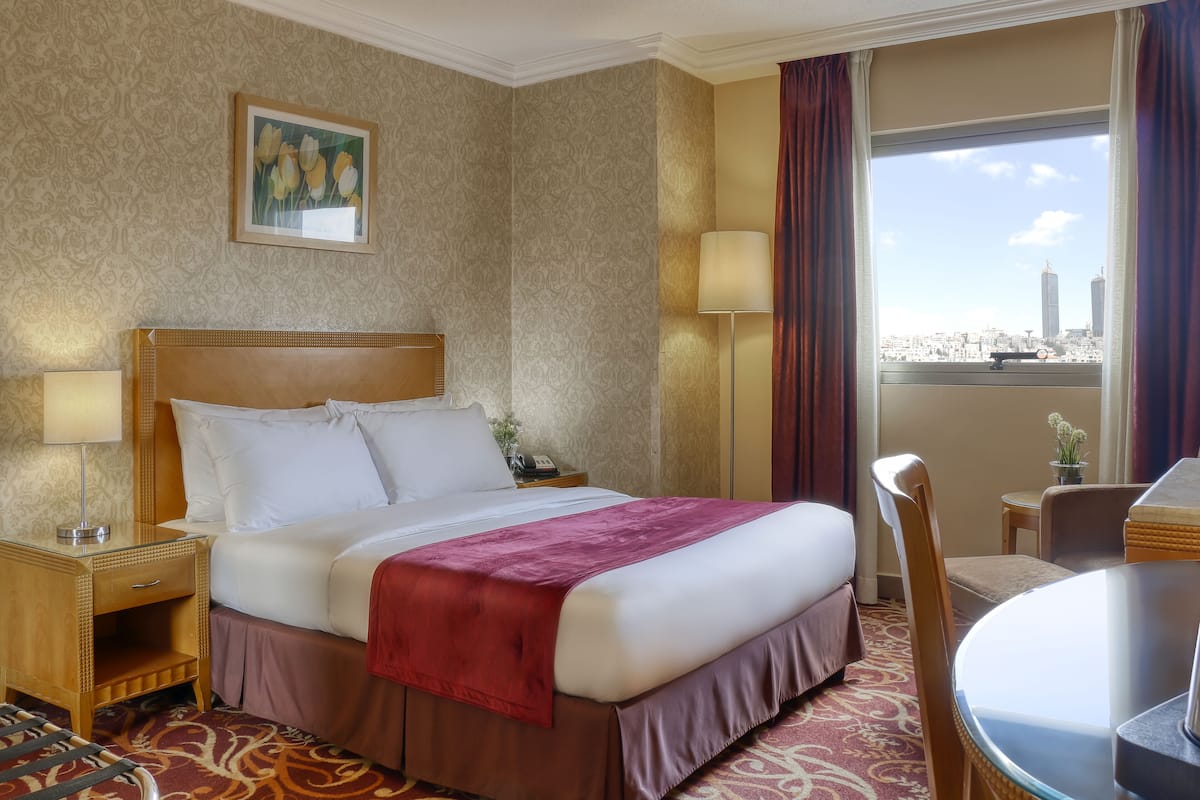 A hotel room with a large bed, a desk, a chair, and a view of the cityscape through the window.