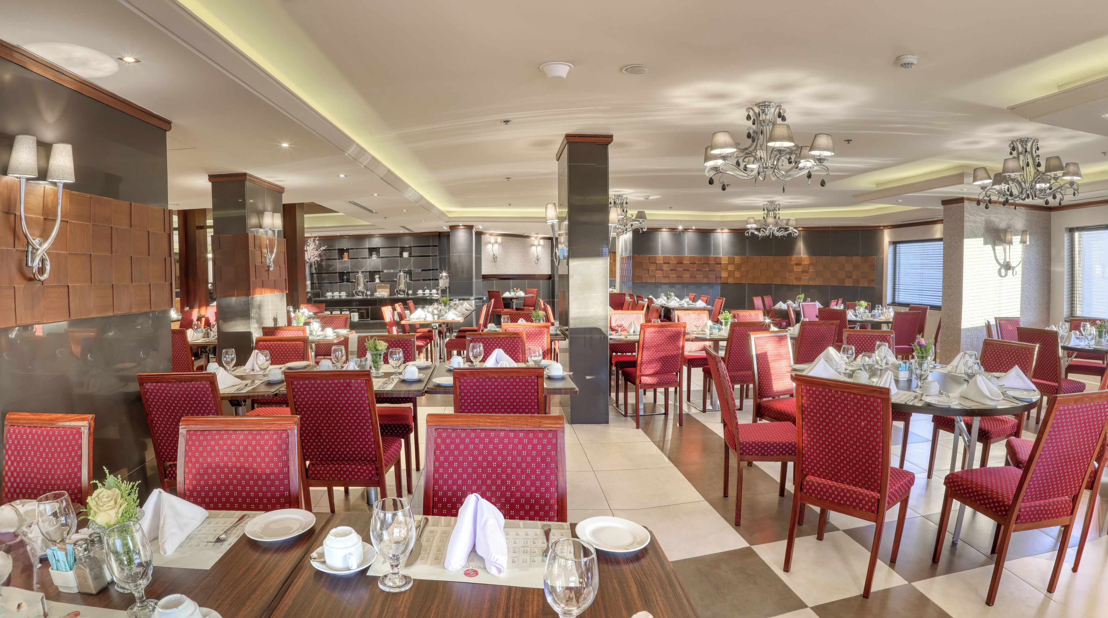 Un comedor de hotel con varias mesas dispuestas para huéspedes, sillas tapizadas en rojo y una lámpara de araña moderna.