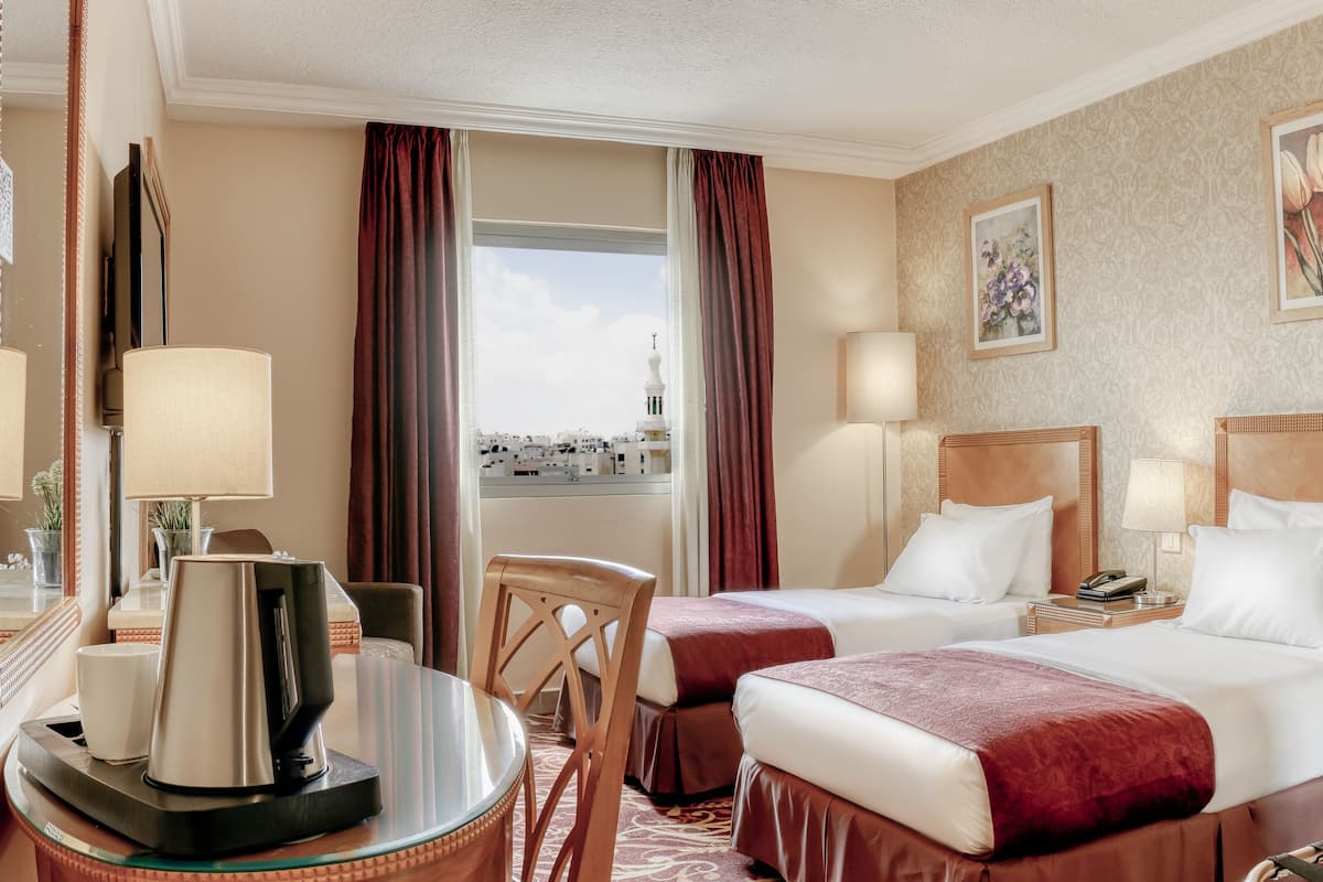 A hotel room with two beds, a desk with a coffee maker, a chair, and a view of the cityscape.