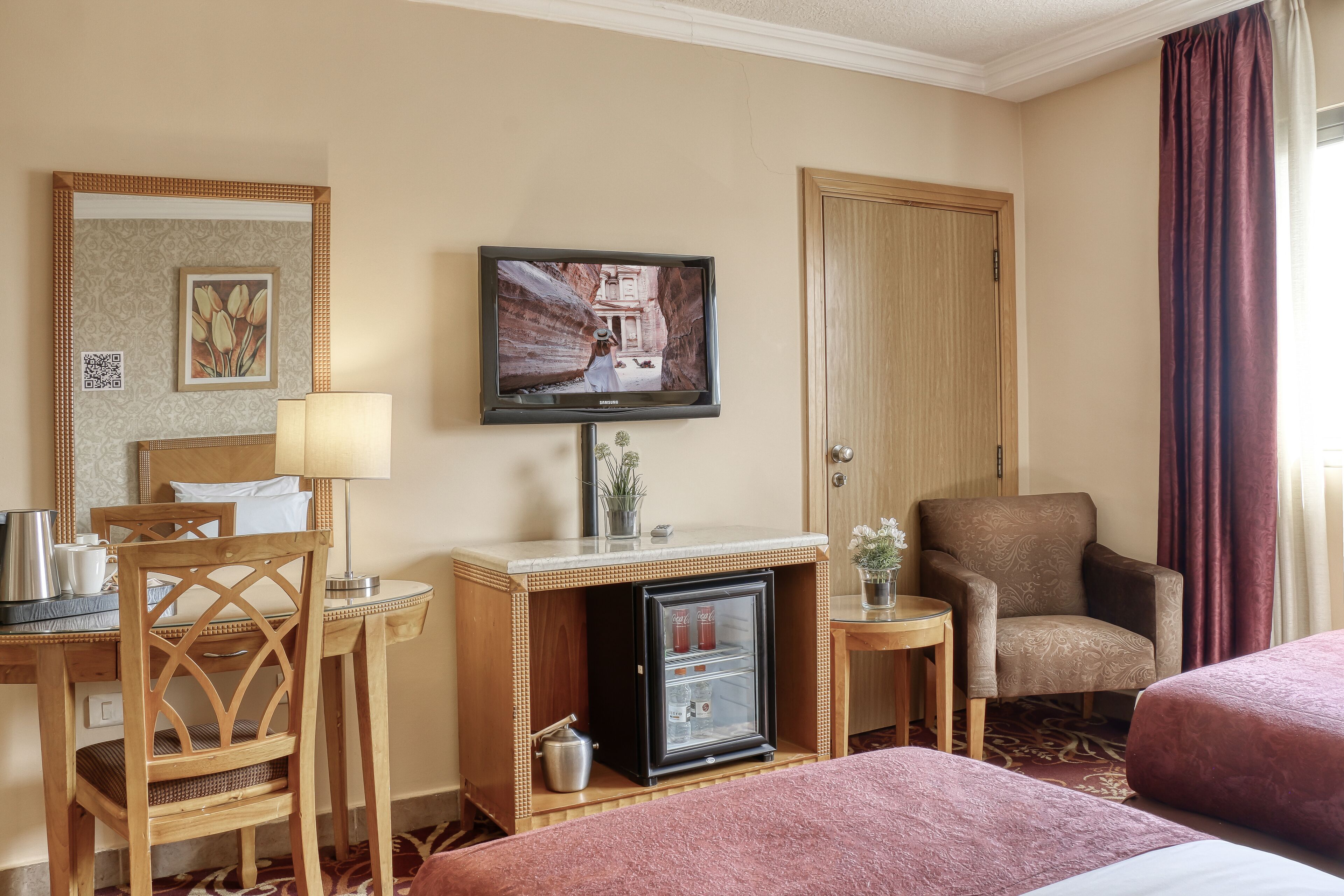 A hotel room with a bed, a desk with a lamp, a television, a refrigerator, and a chair.