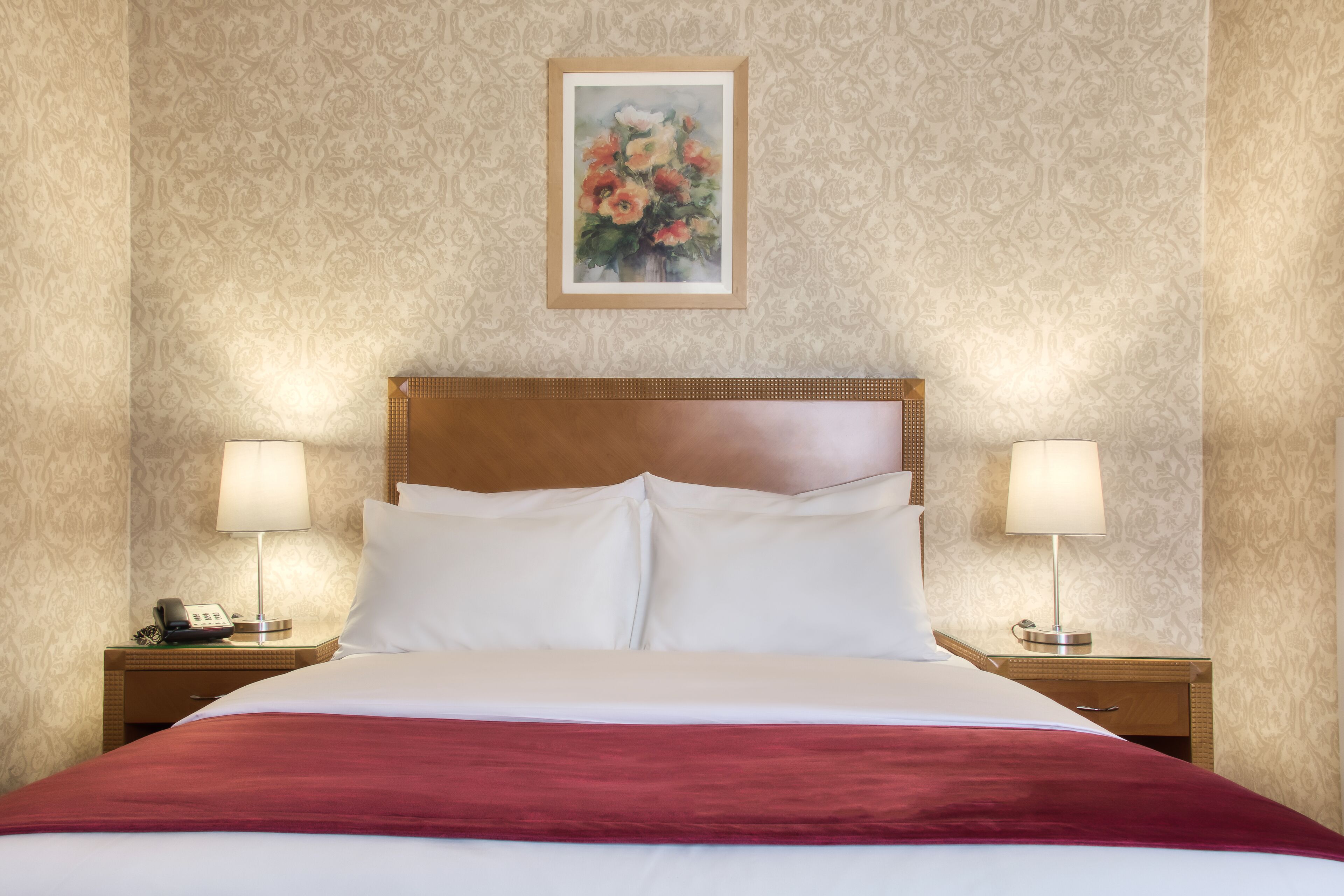 A hotel room with a large bed, two bedside tables with lamps, a telephone, and a framed floral artwork on the wall.