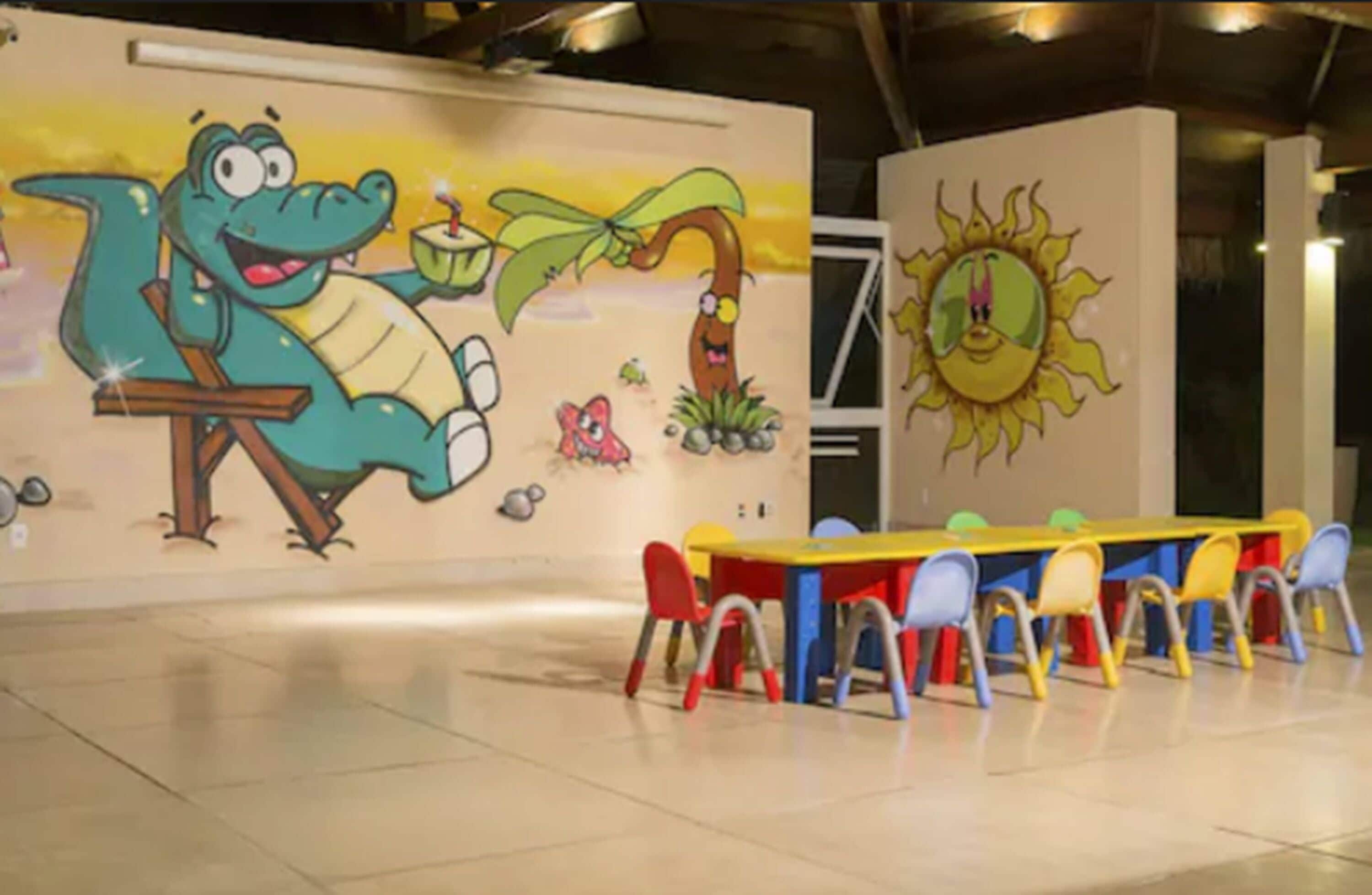 A children's play area with a mural, a long table with colorful chairs, and a sun decoration on the wall.