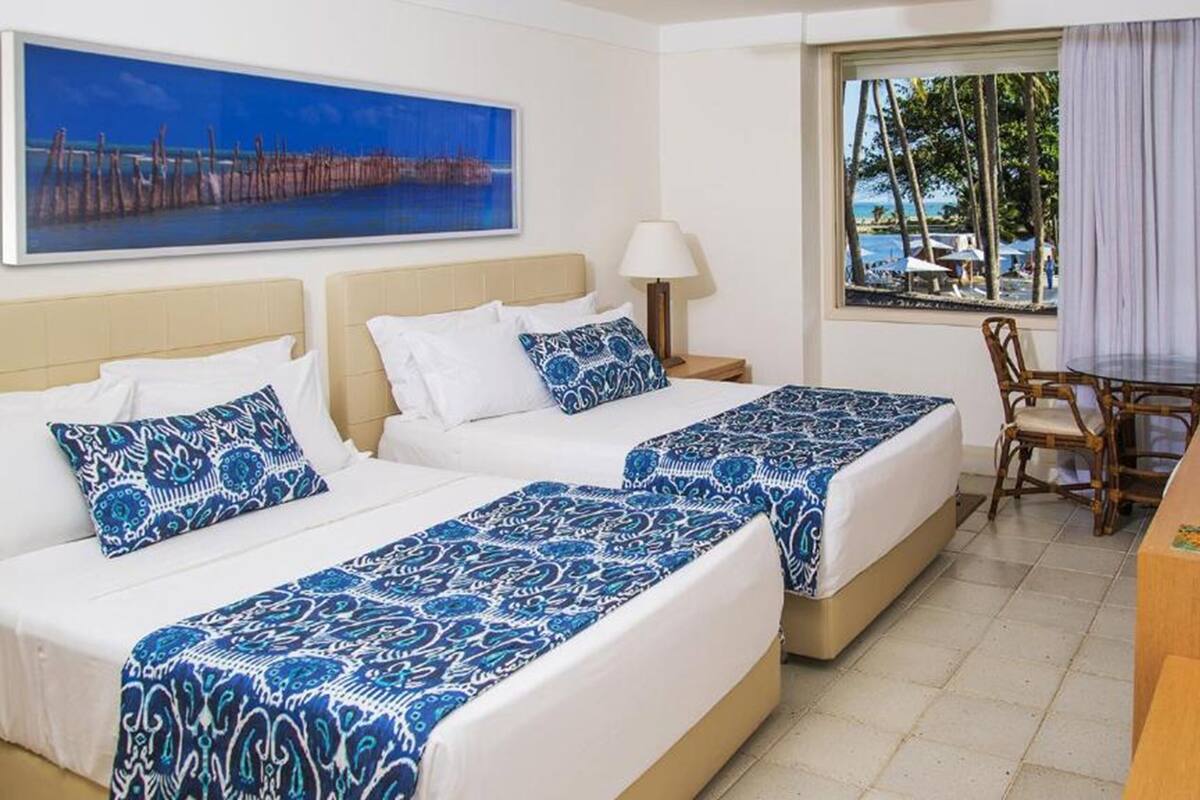 Habitación de hotel con dos camas, un escritorio y ventana con vista a la playa y palmeras.