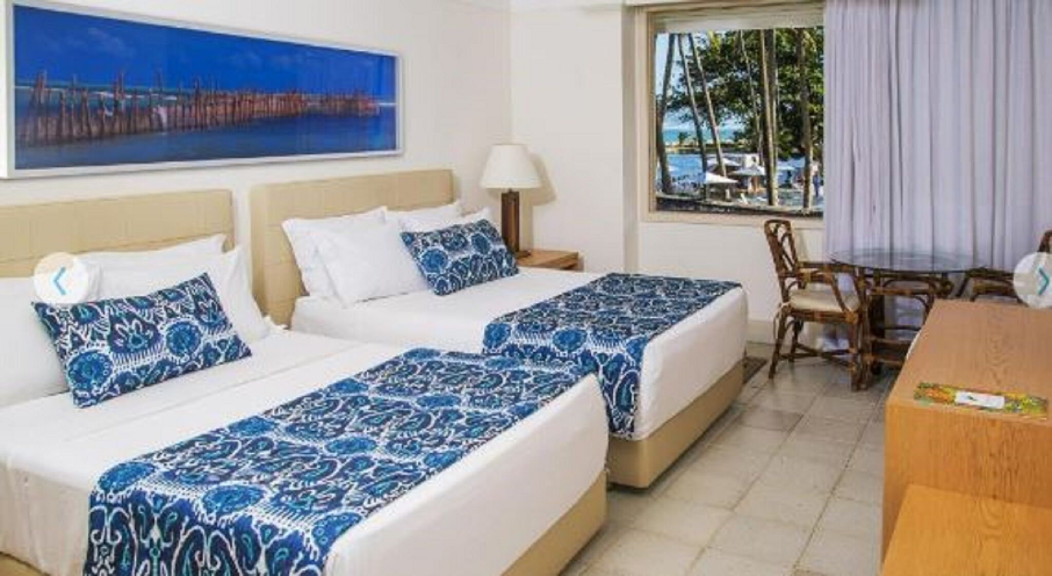 A hotel room with two beds, a desk, a chair, a window with a view of the beach, and a painting on the wall.