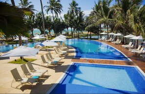 Área de piscina com espreguiçadeiras e guarda-sóis, cercada por palmeiras.
