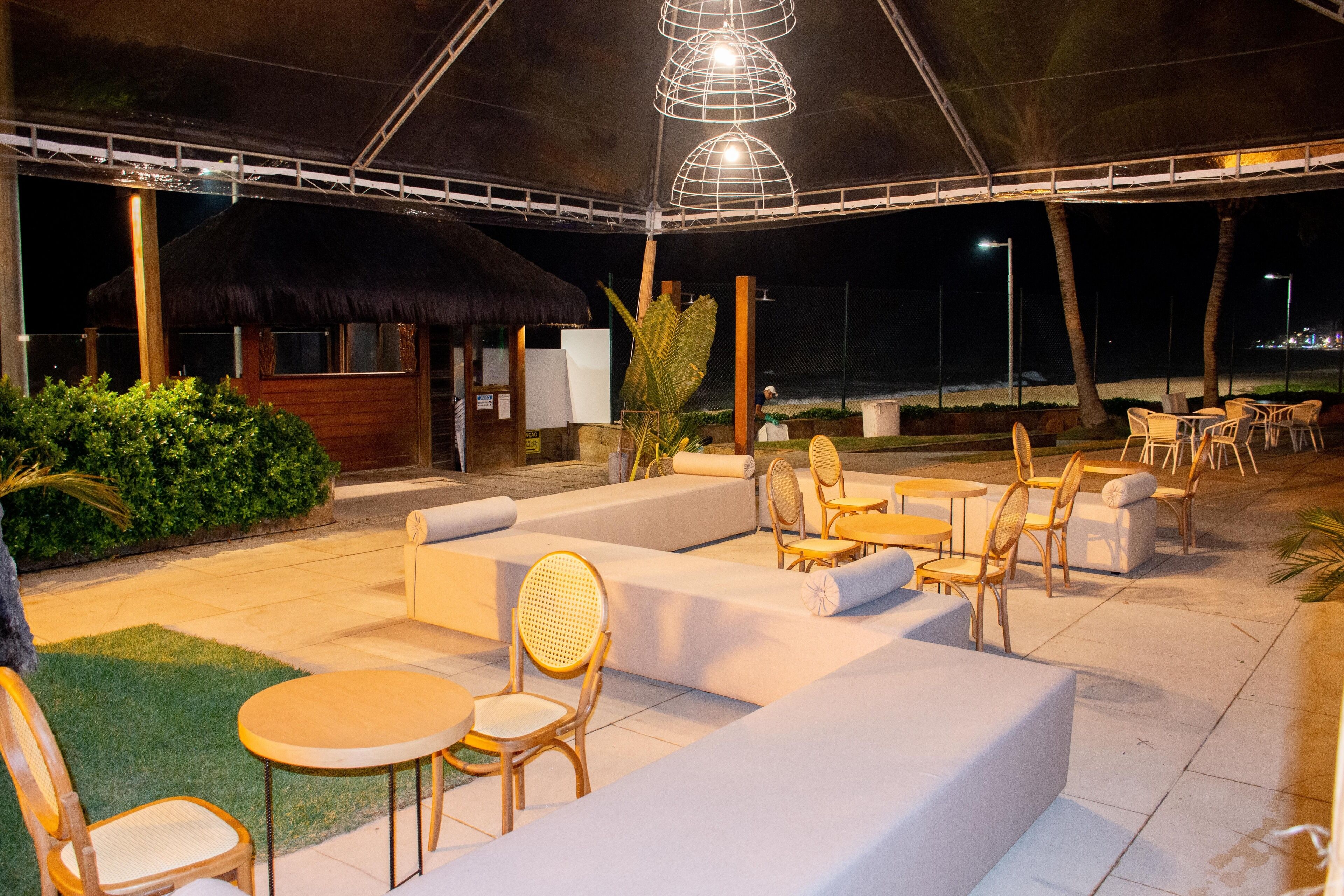 Un área de estar al aire libre con sofás acolchados y sillas de madera, una estructura de techo de paja y un ambiente bien iluminado.
