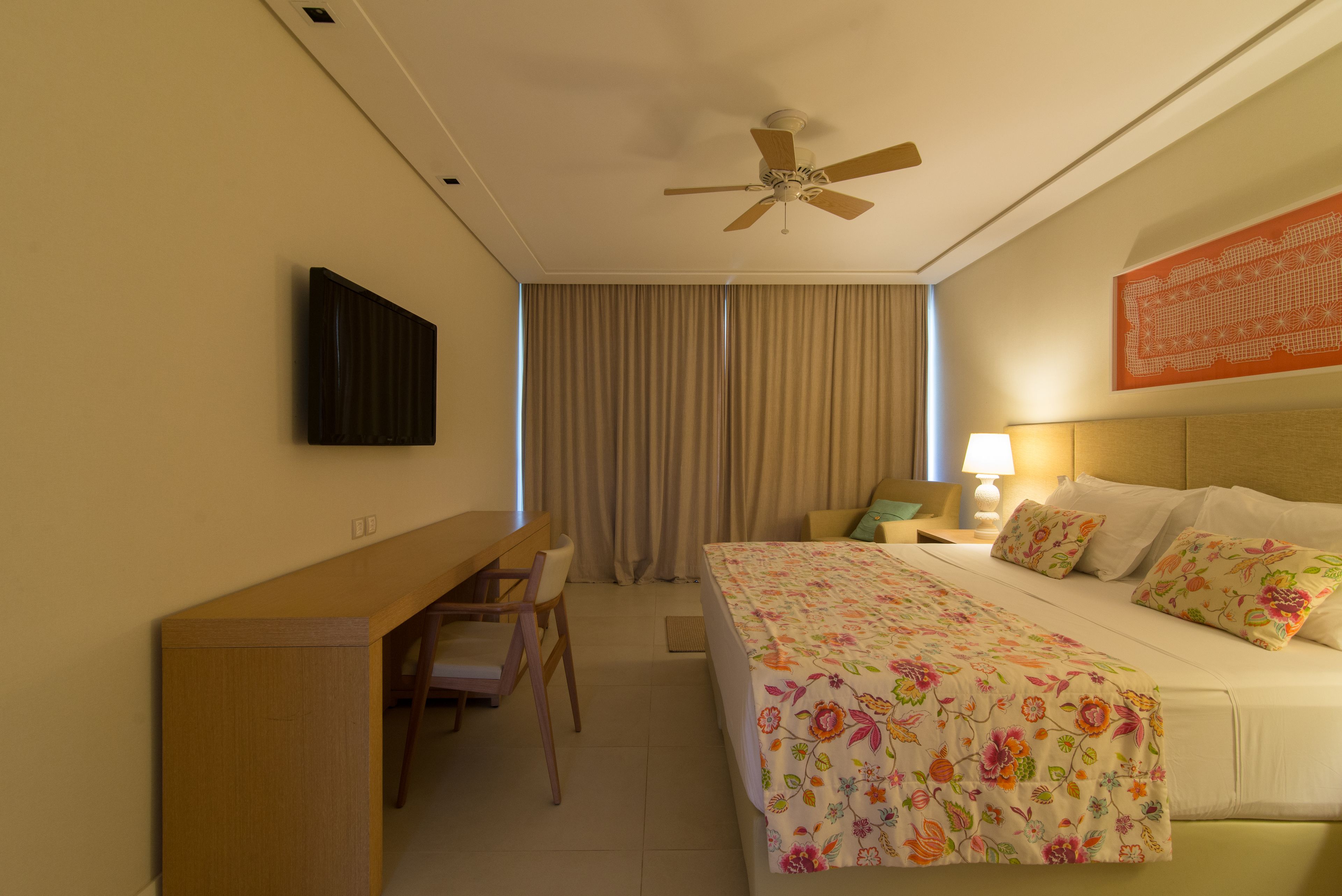 Habitación de hotel con cama, escritorio, silla, ventilador de techo, televisor y ventana con cortinas.
