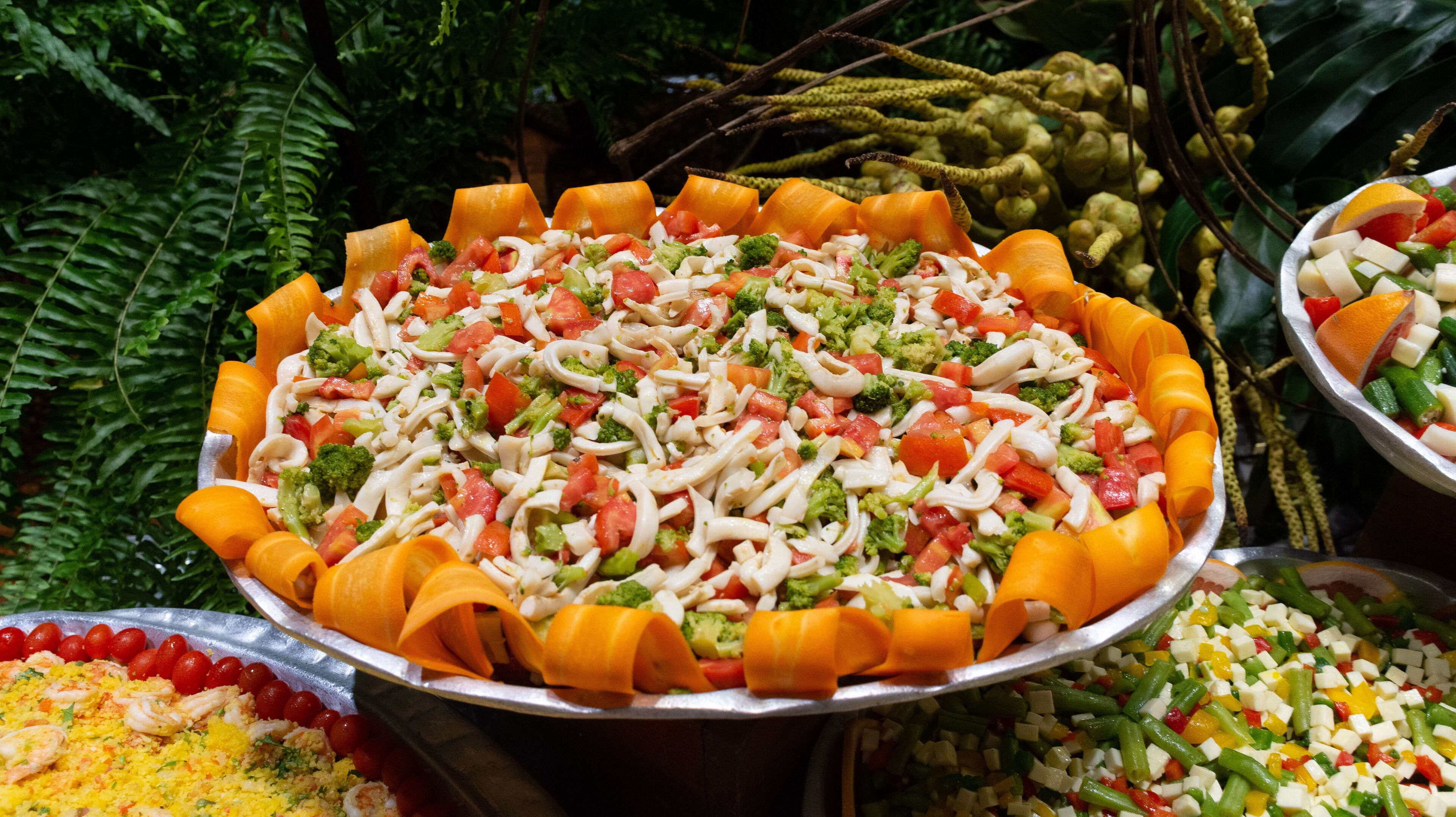 A large platter of mixed vegetables and pasta salad surrounded by other dishes.