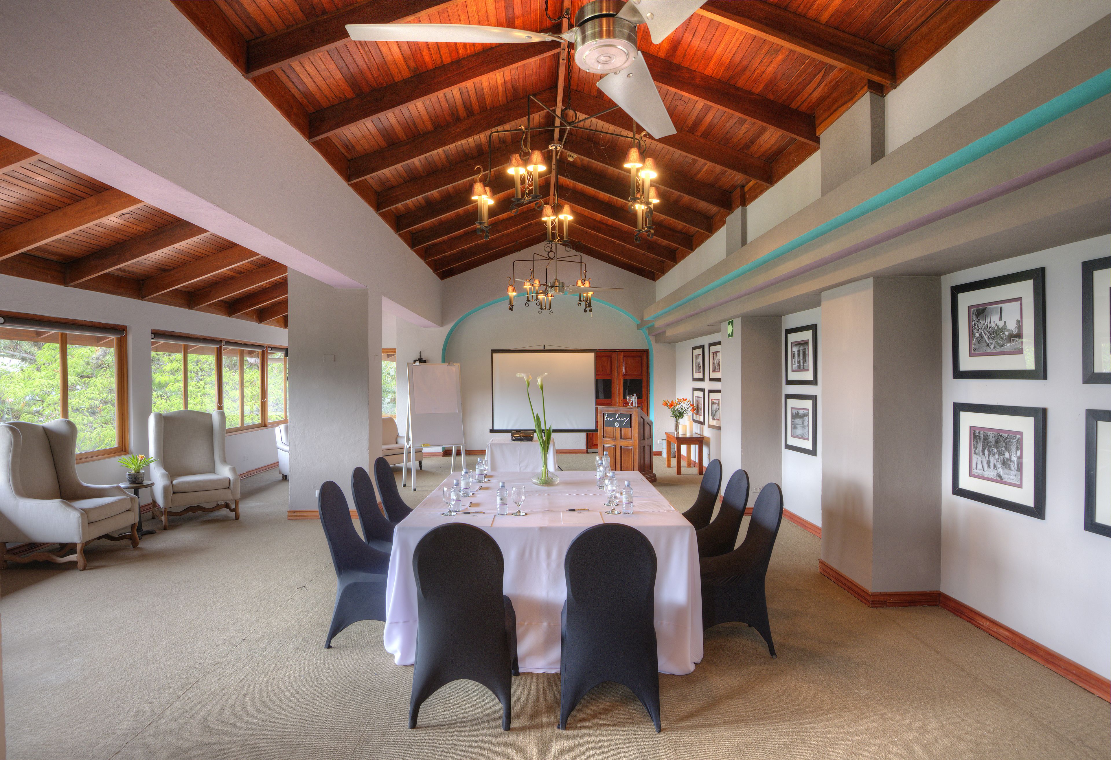 A spacious room with a high ceiling, wooden beams, a chandelier, a dining table set for six, and framed pictures on the walls.