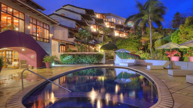 A swimming pool with a clear view of the surrounding architecture, including a multi-story building with balconies and a landscaped garden with palm trees. - Alta Las Palomas Hotel  (Santa Ana)