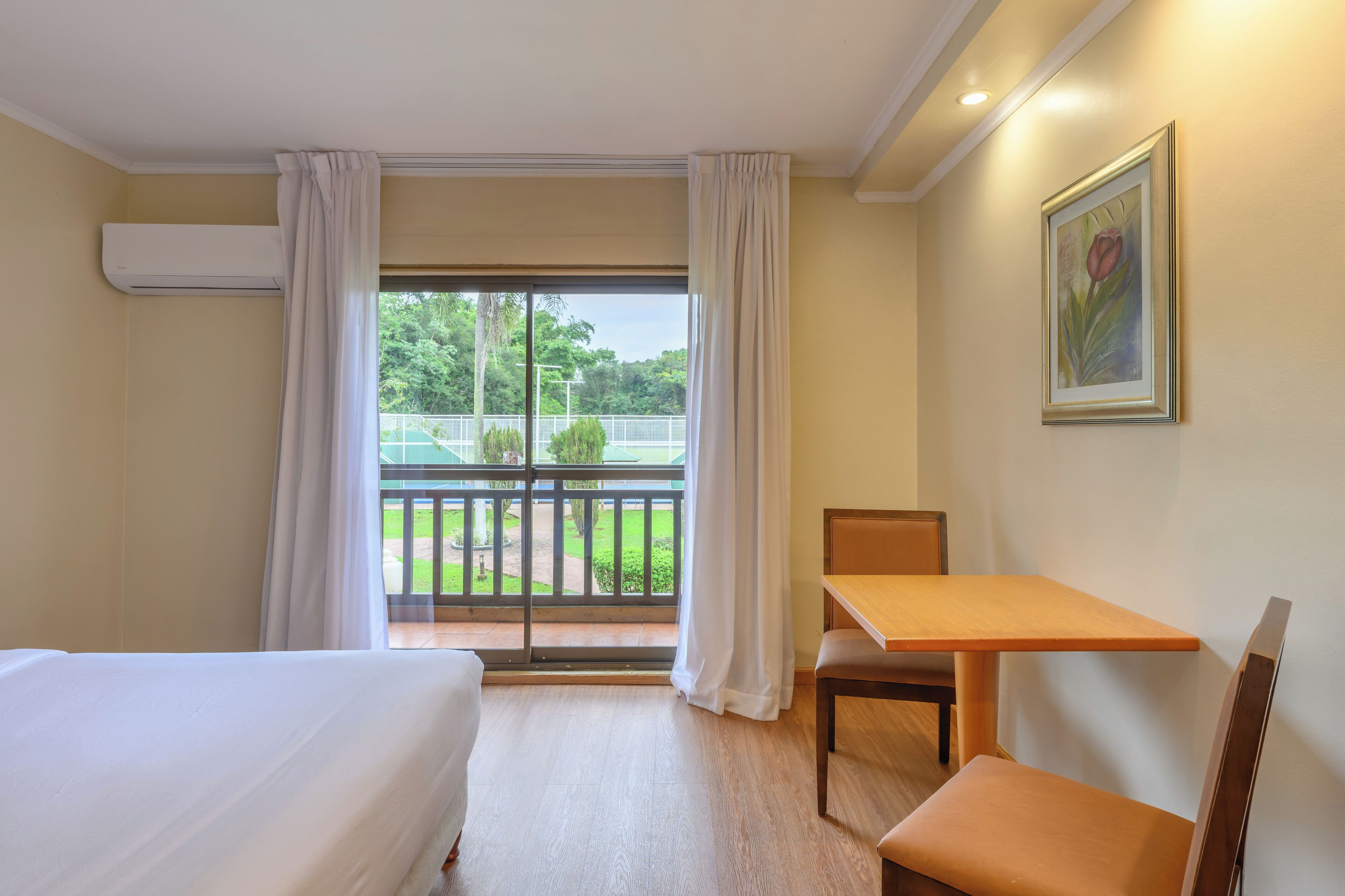 A hotel room with a bed, a desk, a chair, a window with curtains, an air conditioning unit, and a framed picture on the wall.