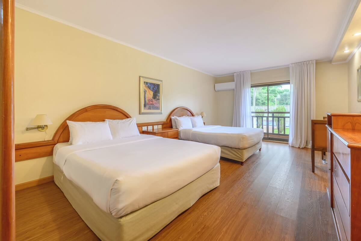 A hotel room with two beds, a desk, and a balcony with curtains.