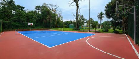 An outdoor basketball court with a blue playing area and red surrounding surface, surrounded by greenery and a fence.