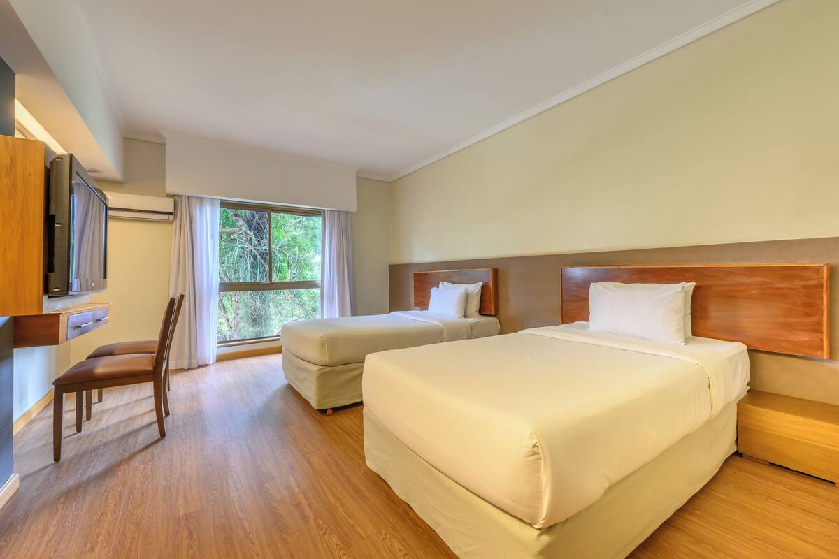 A hotel room with two beds, a wooden floor, a TV, and a window with curtains.