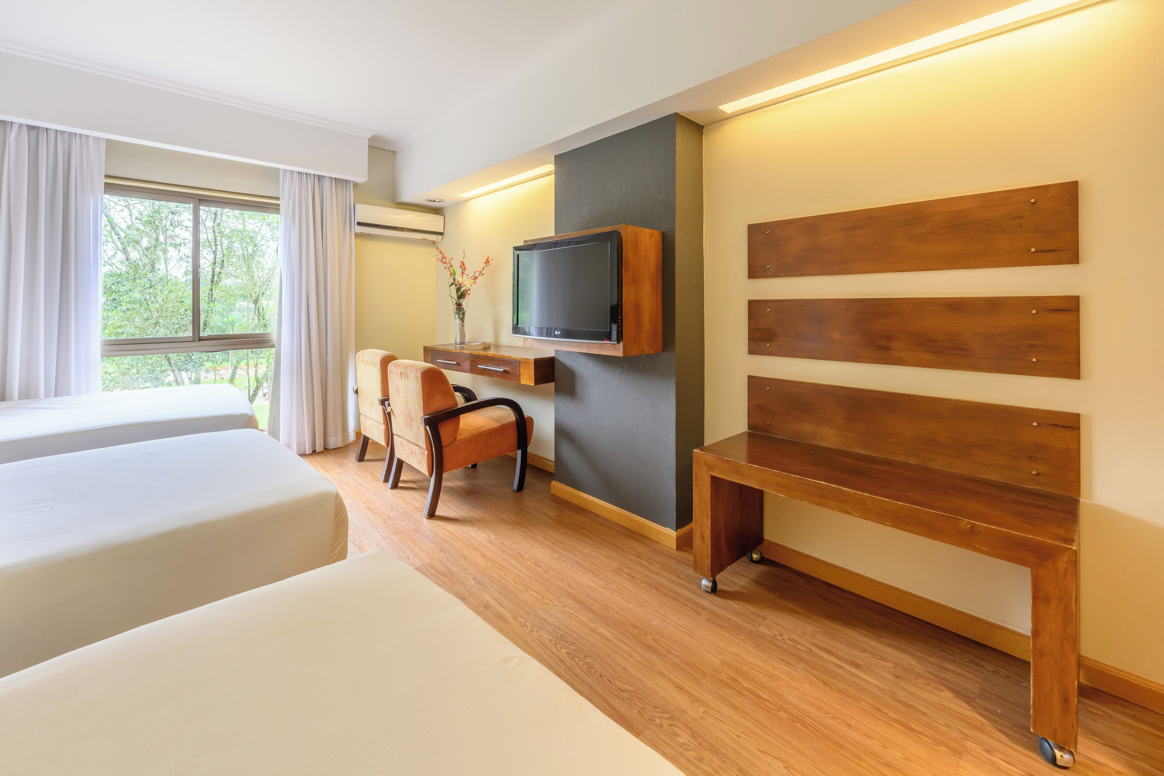 A hotel room with two beds, a desk with a chair, a TV mounted on the wall, and a large window with curtains.