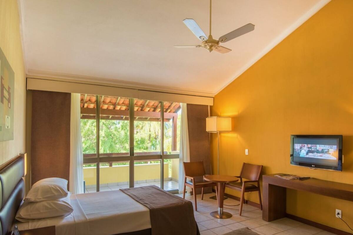 A hotel room with a bed, a desk with a TV, two chairs, a ceiling fan, and a window with a view of greenery.