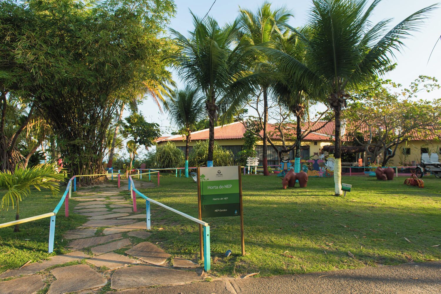 A garden pathway leading to a building with a red roof, surrounded by palm trees and greenery.