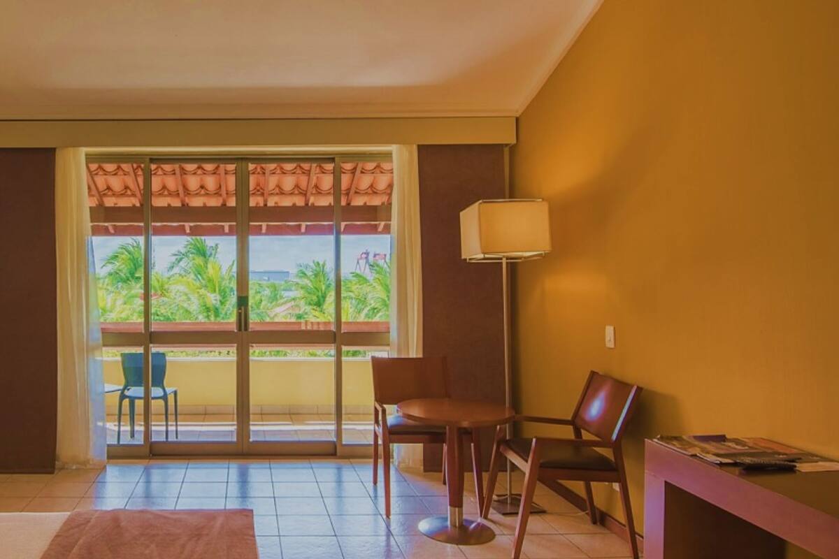 A hotel room with a bed, a desk, a chair, a lamp, and a view of greenery outside.