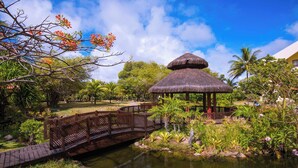 Un pavillon au toit de chaume, un pont en bois et un étang, le tout dans un cadre de jardin tropical.