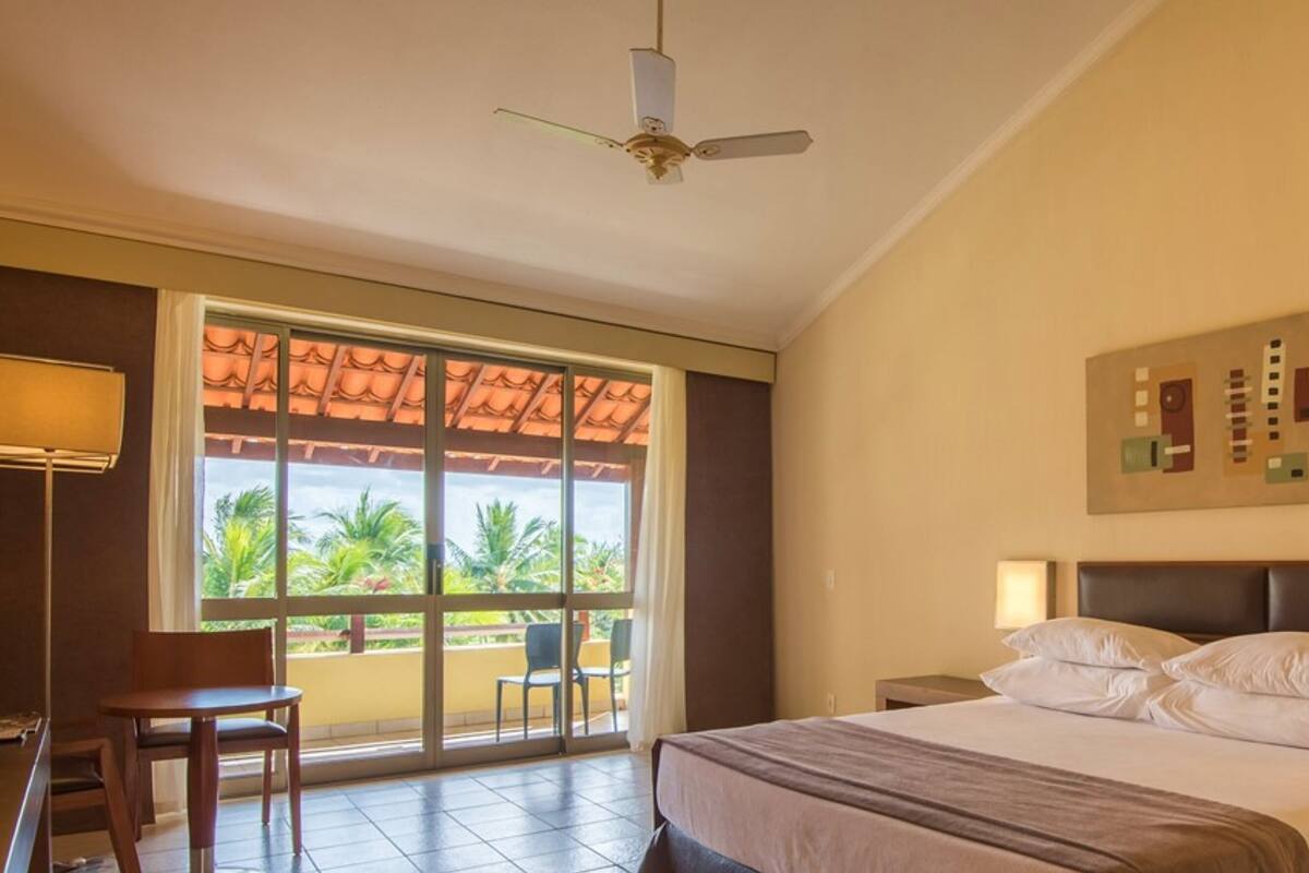 A hotel room with a bed, a desk, a chair, a ceiling fan, a large window with a view of palm trees, and a painting on the wall.