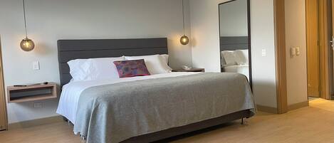 A modern hotel room with a large bed, bedside tables, a nightstand, a bedside lamp, and a mirror on the wall.