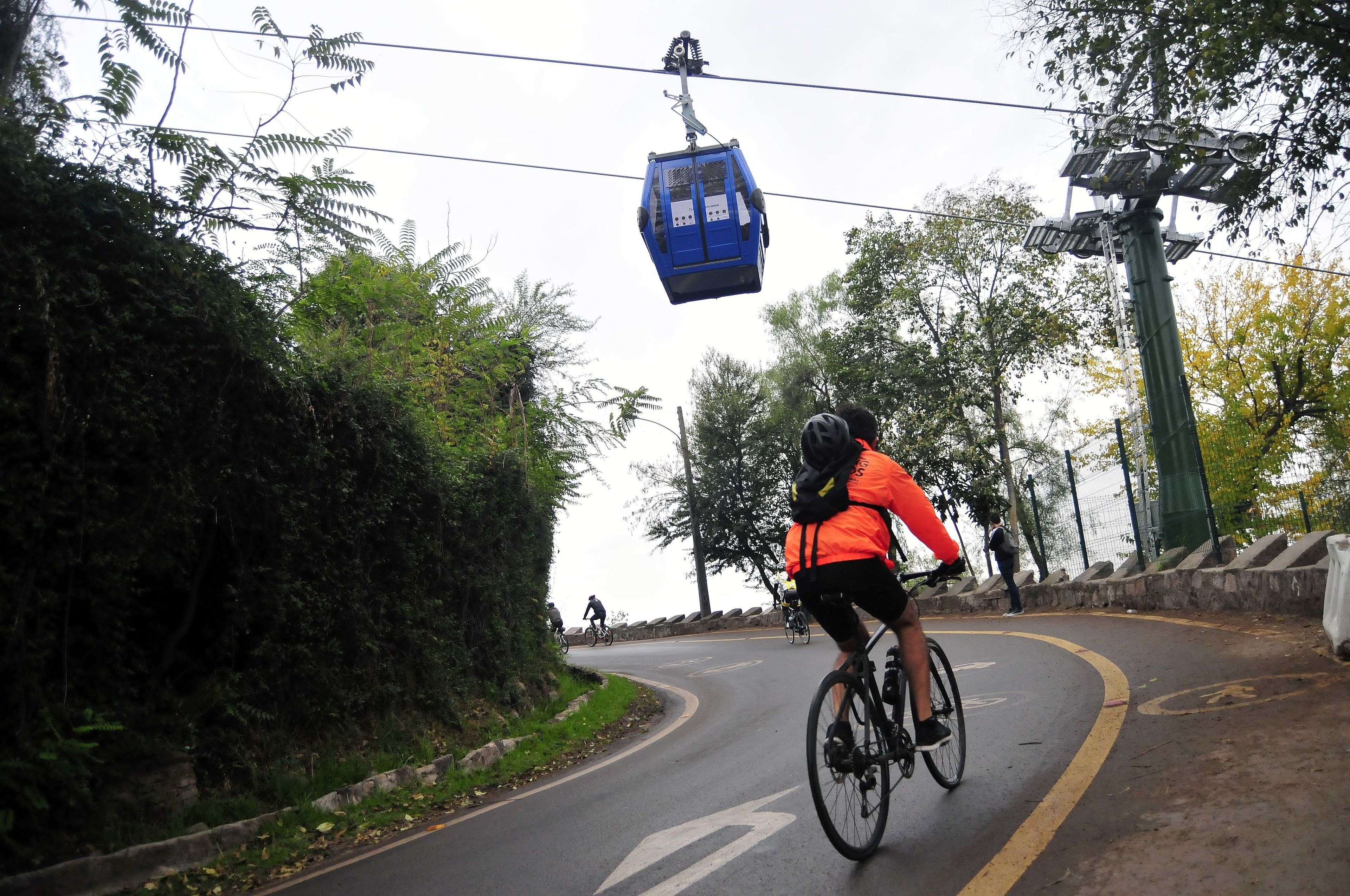 A cyclist on a road with a cable car overhead.