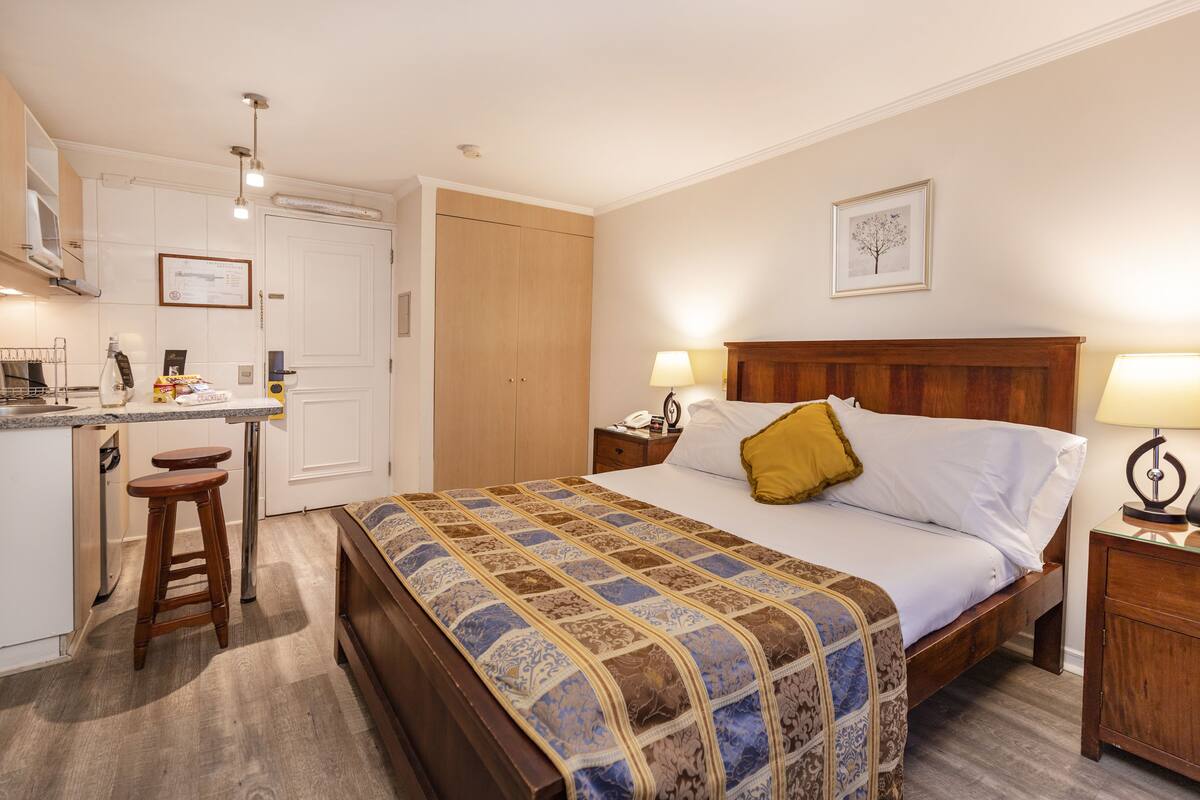 A hotel room with a bed, bedside tables, a desk with a chair, and a kitchen area.