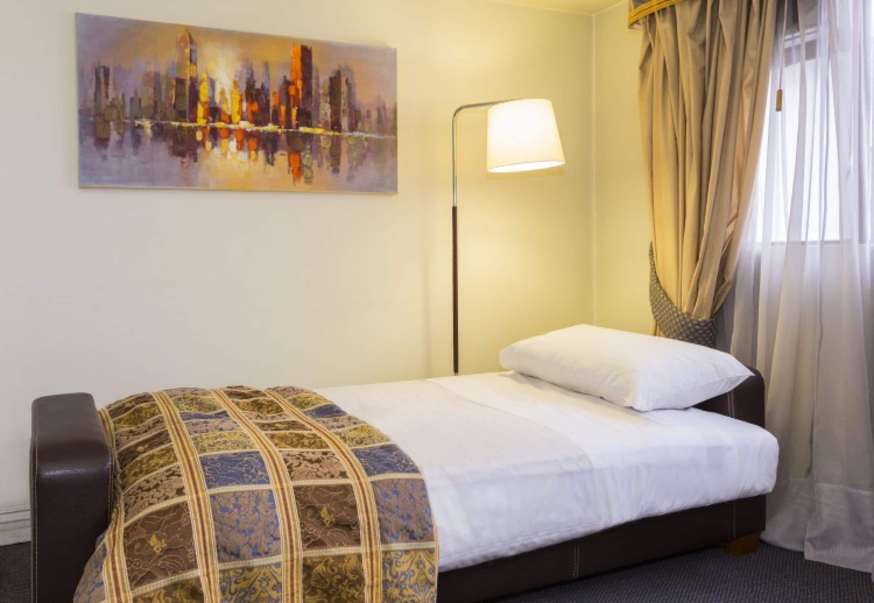 A hotel room with a bed, a painting on the wall, a lamp, and curtains.