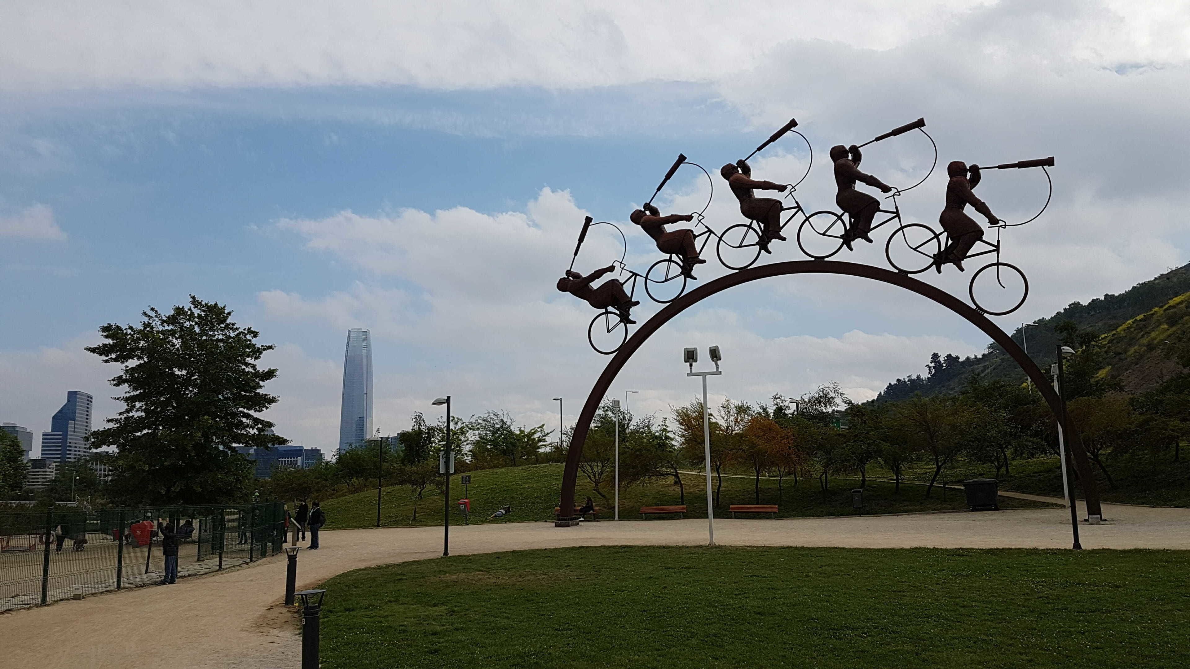 Une sculpture représentant des cyclistes en mouvement, avec en arrière-plan une vue sur les gratte-ciel d’une ville et un parc.