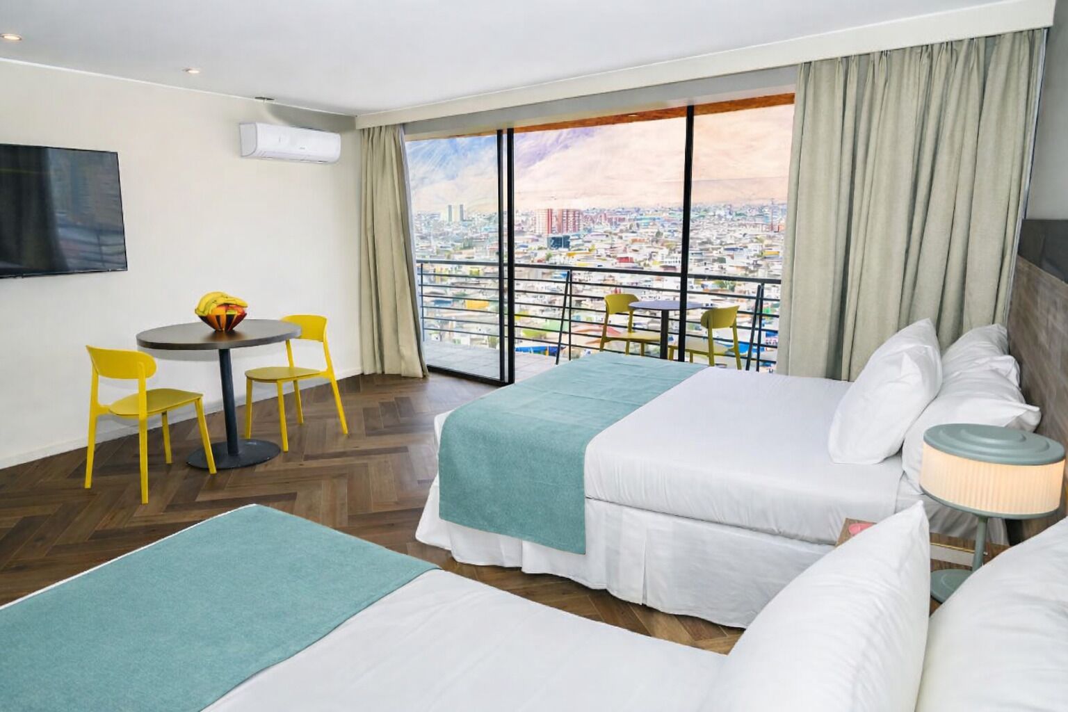 A hotel room with two beds, a dining table with chairs, a TV, and a balcony with a city view.